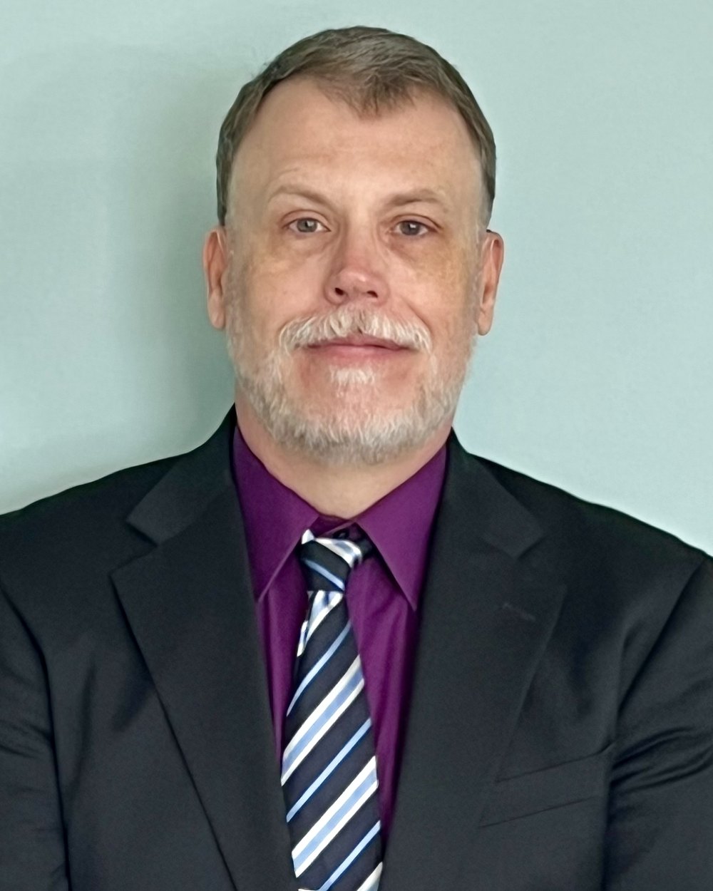 Charles Boswell, wearing a dark suit, purple shirt and striped tie, photographed against a pale wall.